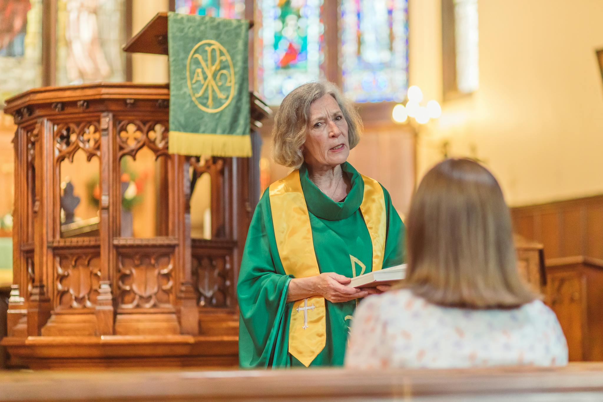 An elderly priest stands in a church pulpit delivering a sermon to a congregation member.